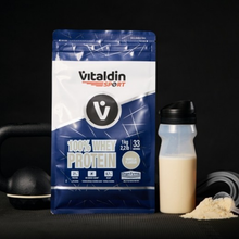 Load image into Gallery viewer, A lifestyle product photograph of the Vitaldin Sport Vanilla Whey Protein bag standing on a gym floor. Next to the bag is a shaker bottle filled with a vanilla-colored protein shake. In the blurred background, gym equipment like dumbbells and weight plates are visible, creating a fitness-oriented atmosphere.