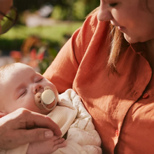 Load image into Gallery viewer, A peaceful baby sleeping while wearing a beige pacifier, held in the arms of a person wearing an orange shirt.