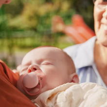 Load image into Gallery viewer, A close-up of a peaceful baby sleeping while wearing a pink pacifier, held in the arms of an older adult.