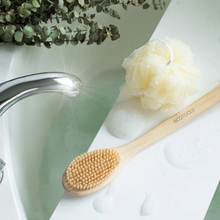 Load image into Gallery viewer, EcoTools bamboo bristle bath brush resting on a bathroom ledge next to a bath shower pouf and soapy lather.