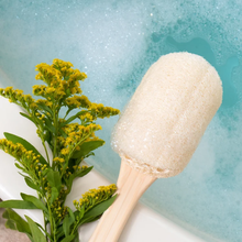 Load image into Gallery viewer, EcoTools loofah bath brush resting on the edge of a bubble bath next to bright yellow flowers.