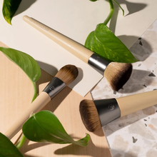 Load image into Gallery viewer, EcoTools makeup brushes laid out on a beige surface surrounded by green plant leaves.