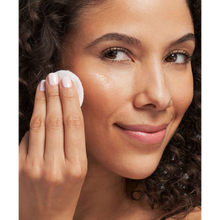 Load image into Gallery viewer, A close-up lifestyle image of a smiling person with curly hair using a cotton pad to apply NeoStrata Oily Skin Solution to their cheek.