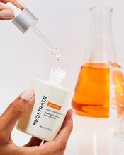 Load image into Gallery viewer, Hand holding Neostrata ENLIGHTEN Dual Acid Brightening Peel bottle with a dropper dispensing the liquid; a beaker and flask are in the background, suggesting a scientific skincare product.