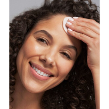 Load image into Gallery viewer, A close-up lifestyle image of a person with curly hair smiling while using a circular white NeoStrata PHA Renewal Pad on their forehead. The person has a light-colored manicure and clear, glowing skin.