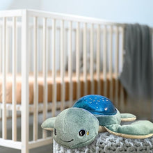 Load image into Gallery viewer, The Pabobo Aqua Dream Turtle ocean soother placed in the foreground of a nursery setting, near a clean, white wooden crib.
