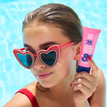 Load image into Gallery viewer, A woman wearing heart-shaped sunglasses by a pool holding the A.N.D. beauty Clear Sun Balm.
