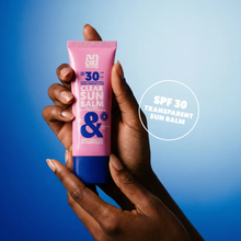 Load image into Gallery viewer, Hands holding the A.N.D. beauty Clear Sun Balm tube against a blue background, highlighting the transparent formula.