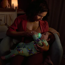 Load image into Gallery viewer, A woman in a red shirt gently feeds a baby in a dimly lit room. The baby wears colorful toucan-patterned pajamas and drinks from a bottle with a glowing green ring.