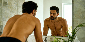 A shirtless man gazes at his reflection in the mirror, examining his appearance with a thoughtful expression.