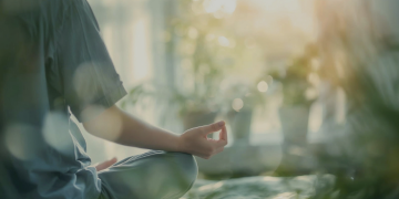 Person sitting cross-legged in a peaceful indoor setting, practicing meditation with one hand resting in a mindfulness pose, surrounded by soft natural light and blurred greenery. 
