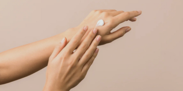 Close-up of hands applying moisturizing cream to the back of one hand against a soft beige background, highlighting smooth skin and nail care.