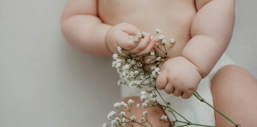 Close-up of a baby wearing a white diaper, holding delicate white flowers in both hands against a soft neutral background.