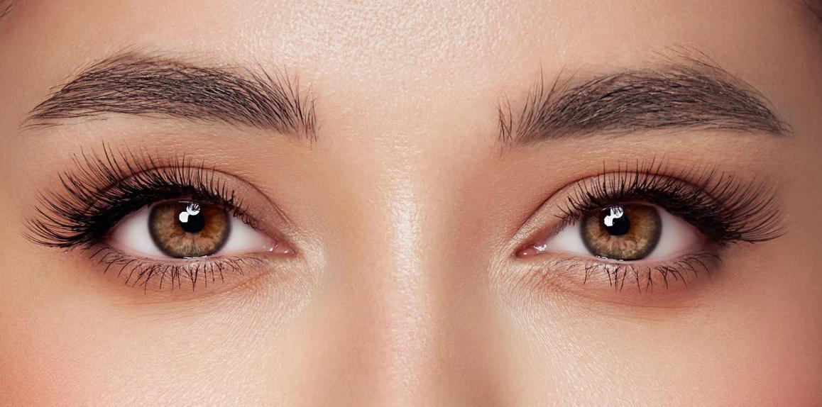 Close-up of a person’s brown eyes with long curled eyelashes, defined dark eyebrows, and smooth skin under soft lighting.