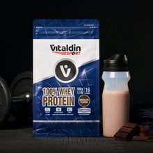 Load image into Gallery viewer, A lifestyle product photo featuring a bag of Vitaldin Sport Chocolate Whey Protein standing next to a shaker bottle filled with a chocolate protein shake. In the background, a weight plate is visible on the left, and pieces of dark chocolate are arranged in the foreground on the right.
