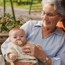 Load image into Gallery viewer, An older woman smiling while holding a baby who is wearing a beige pacifier.
