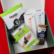 Load image into Gallery viewer, An open Botika health and sports subscription box on a red background, featuring Vitaldin Sport Power Gummies, Biofrost Active and Relief gels, a Vitaldin protein bar, and a Uriel wrist support brace.

