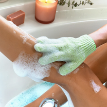 Load image into Gallery viewer, Person using a sudsy green EcoTools shower glove to exfoliate their leg in a bath.
