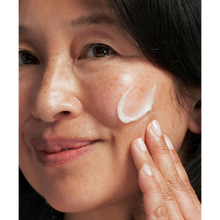 Load image into Gallery viewer, A smiling person applying Neostrata Enlighten Skin Brightener cream to their cheek, highlighting its moisturizing and brightening effects.
