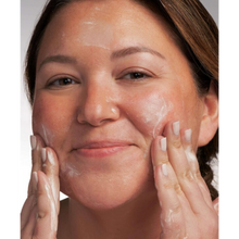 Load image into Gallery viewer, A close-up lifestyle image of a person with a slight smile massaging the white, lathering facial cleanser into their cheeks with their fingertips.
