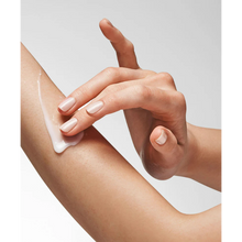 Load image into Gallery viewer, A close-up lifestyle image of a person applying a white, creamy lotion to their arm. The person’s fingers, featuring a light neutral manicure, are smoothing the product onto the skin against a clean, light grey background.
