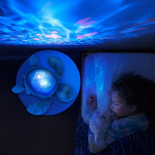 Load image into Gallery viewer, Overhead view of a child sleeping peacefully, illuminated by the Pabobo Aqua Dream Turtle projecting a calming ocean ripple pattern on the ceiling and walls.
