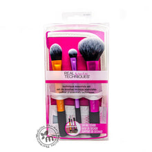 Load image into Gallery viewer, A set of Real Techniques travel essentials makeup brushes packaged in a clear plastic case with pink and white labeling, including brushes for foundation, concealer, and sea salt spray.
