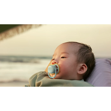 Load image into Gallery viewer, A close-up of an infant using a mint green Philips Avent Ultra Air soother while resting on a beach at sunset.
