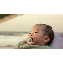 Load image into Gallery viewer, A close-up of an infant using a butterfly-themed Philips Avent Ultra Air soother while resting on a beach at sunset.

