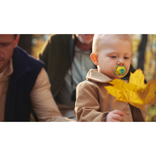 Load image into Gallery viewer, A toddler focused on an autumn leaf while using a yellow and green Philips Avent Ultra Air frog soother during an outdoor family trip.
