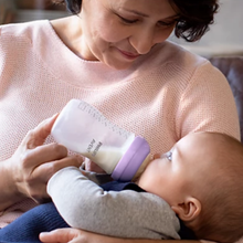 Load image into Gallery viewer, A woman in a pink sweater looks down lovingly at a baby while feeding them with a Philips Avent bottle featuring a purple cap.
