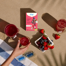 Load image into Gallery viewer, Top-down view of Yogi Frutos del Bosque cold brew tea box on the sand with glasses of red iced tea, a bowl of fresh berries, and sunglasses.
