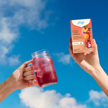 Load image into Gallery viewer, Two hands raised against a bright blue sky with clouds, one holding a glass mason jar mug filled with red iced tea and the other holding a box of Yogi Erdbeer-Orange cold brew tea.
