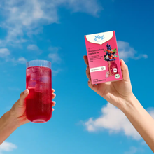 Load image into Gallery viewer, A hand holding a pink box of Yogi Cold Brew Forest Fruit tea next to a tall glass of iced red berry tea against a vibrant blue sky.
