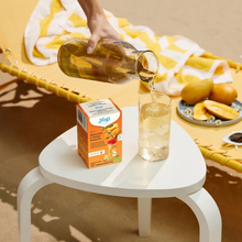 Load image into Gallery viewer, A hand pouring freshly brewed yellow iced tea from a glass carafe into a tall glass with ice next to a box of Yogi Melocoton y Mango on a white table.
