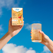 Load image into Gallery viewer, Two hands raised against a bright blue sky with clouds, one holding a box of Yogi Cold Brew Pfirsich-Mango tea and the other holding a tall glass of yellow iced tea with ice.

