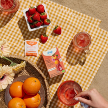 Load image into Gallery viewer, Top-down flat lay view of Yogi Fresa y Naranja tea box on a yellow checkered picnic blanket with fresh oranges, a bowl of strawberries, and mason jars of iced red tea.
